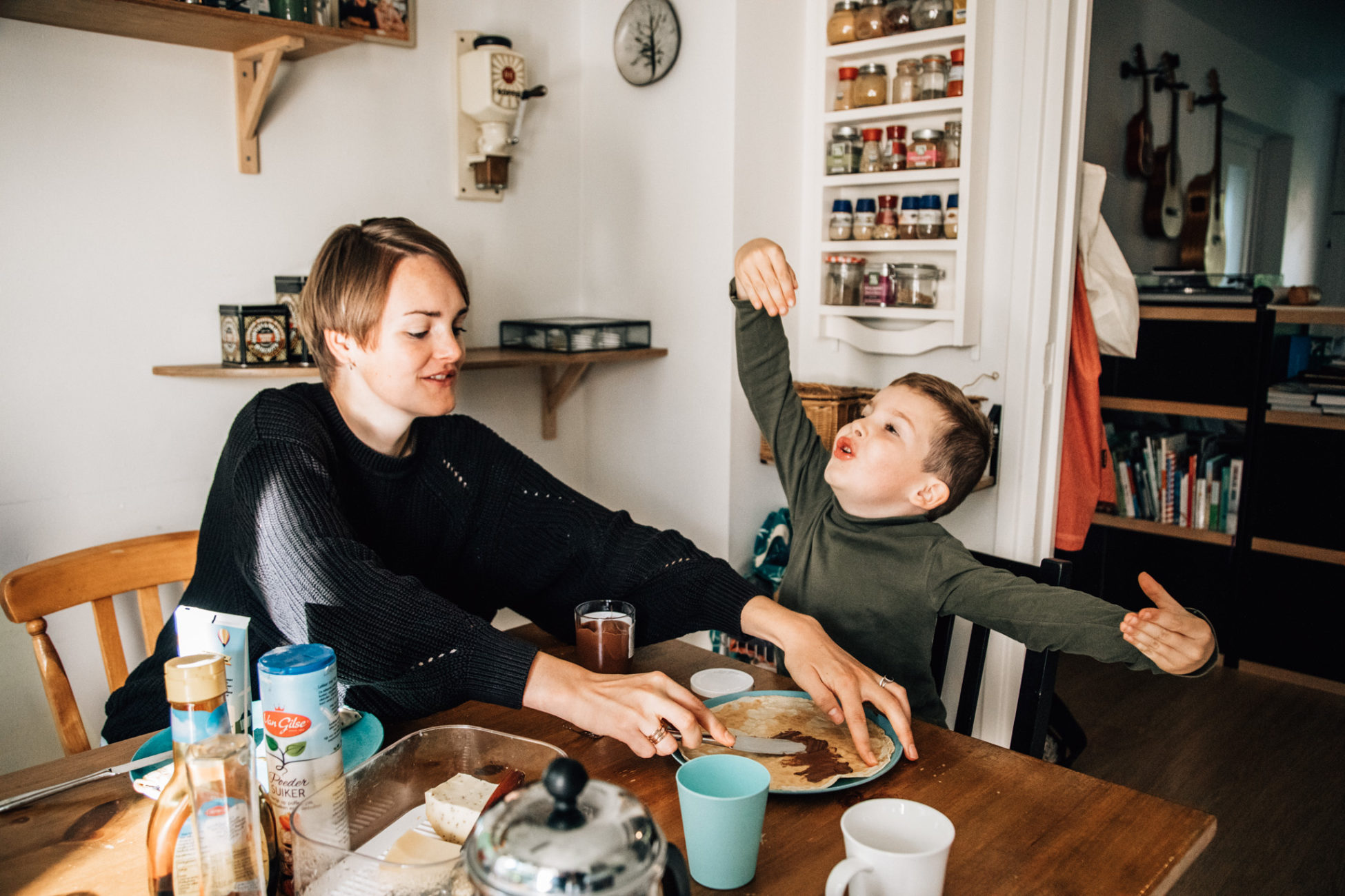 Things you can do during your family photoshoot: to have breakfast together: Mom and child having breakfast together in the kitchen. Kid has arms wide open, like talking of a big explosion while his mom is spreading nutella on his pannenkoek