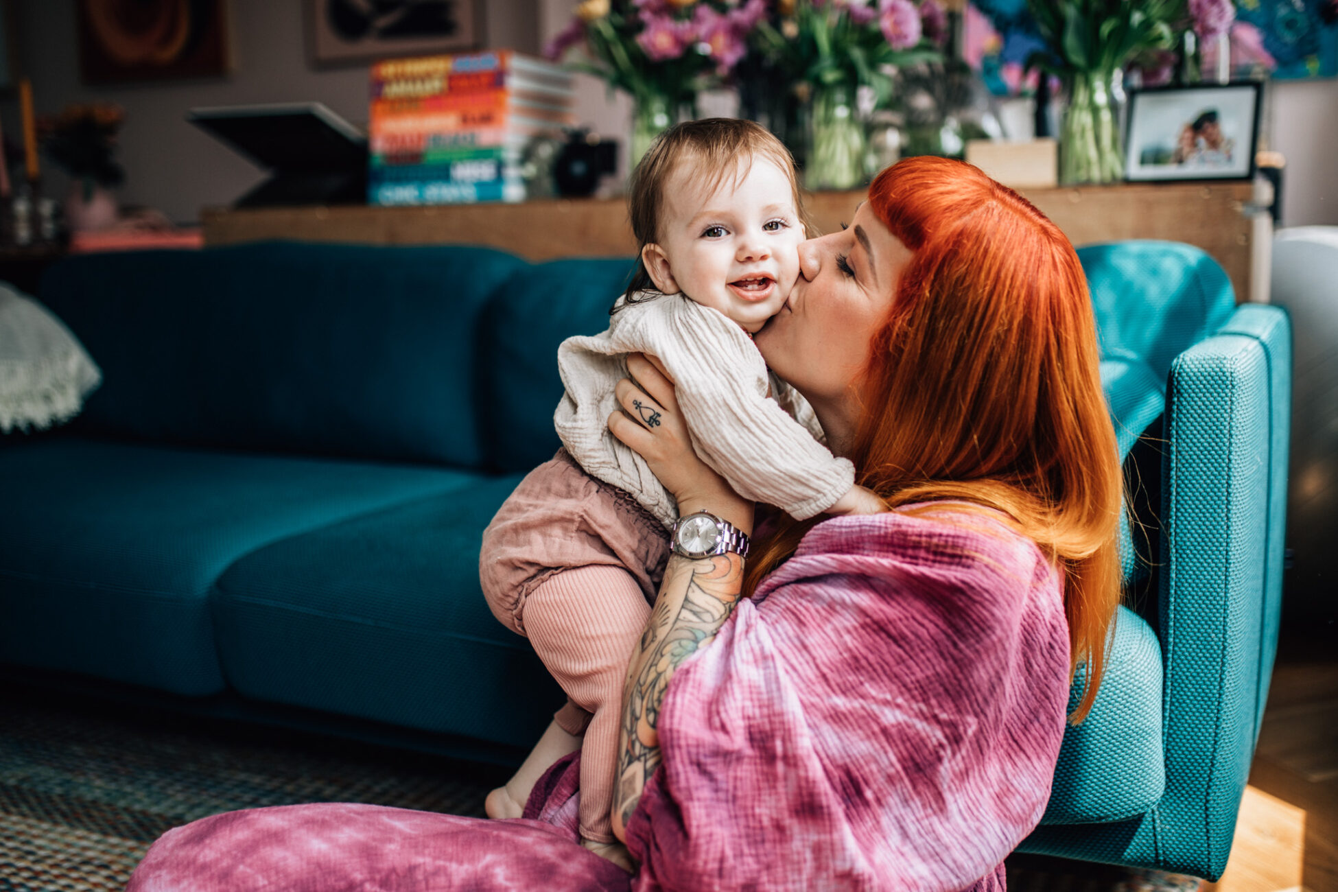 Mom kissing her little girl on her cheek. They're sitting on the floor of their livigroom. Mom is wearing a purple hand dyed dress, she has amazing orange hair and some tattoos on her arm and hand. The photoshoot took place in their home, in Amsterdam.